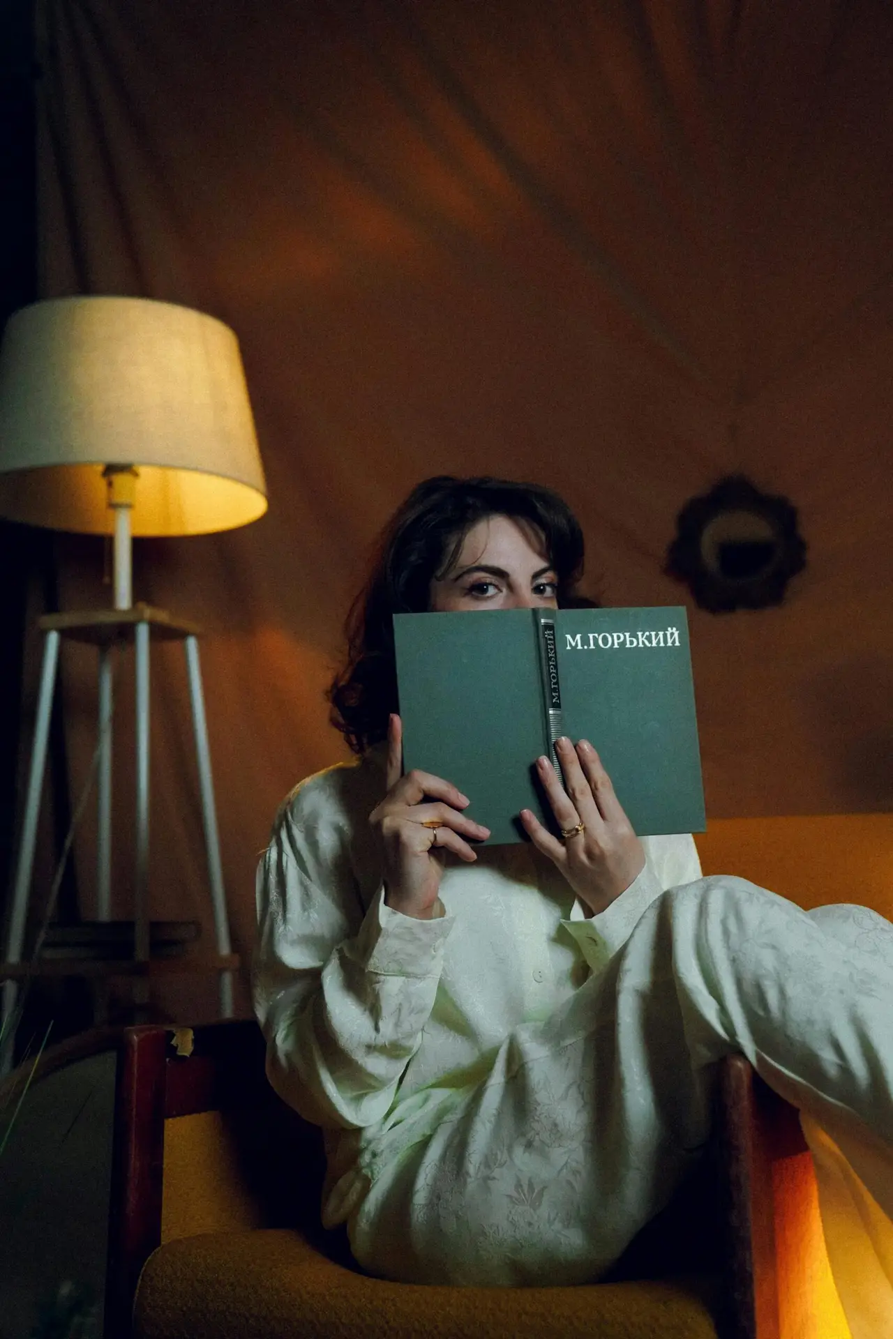 A girl sitting on a couch, on hand a book holding in front of her face which shows only her eyes. Background shows a lamp.