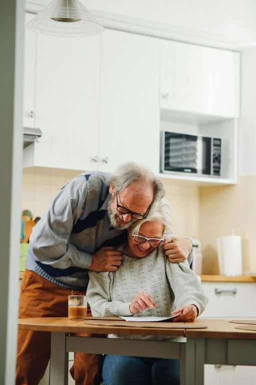 A cheerful elder couple. The male partner is hugging the female and both are looking at something on the table laughing together.