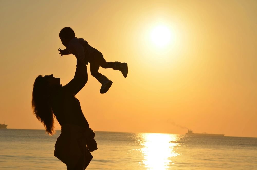 A sea beach with the sunlight. In front of the view a mother is holding her child high and the child is laughing.