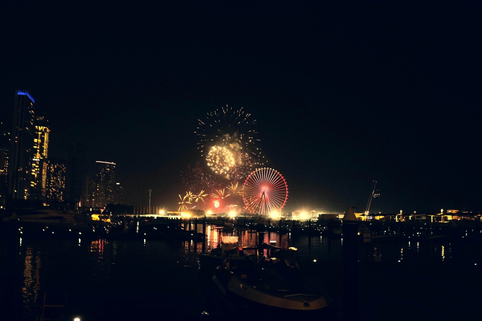 A view of a city by a river showing lights and fireworks in the sky. The shadow of the city is reflecting on the river water.